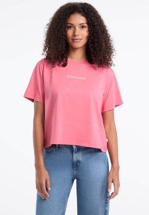 Camiseta rosa corta de mangas cortas, con el logo "GUESS JEANS" en blanco. Combinada con jeans de talle alto de color azul claro. Textura suave.