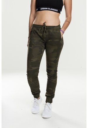 Femme portant un pantalon de jogging camouflage vert foncé, un crop top noir, des baskets blanches et un bracelet violet, debout, une main dans la poche.
