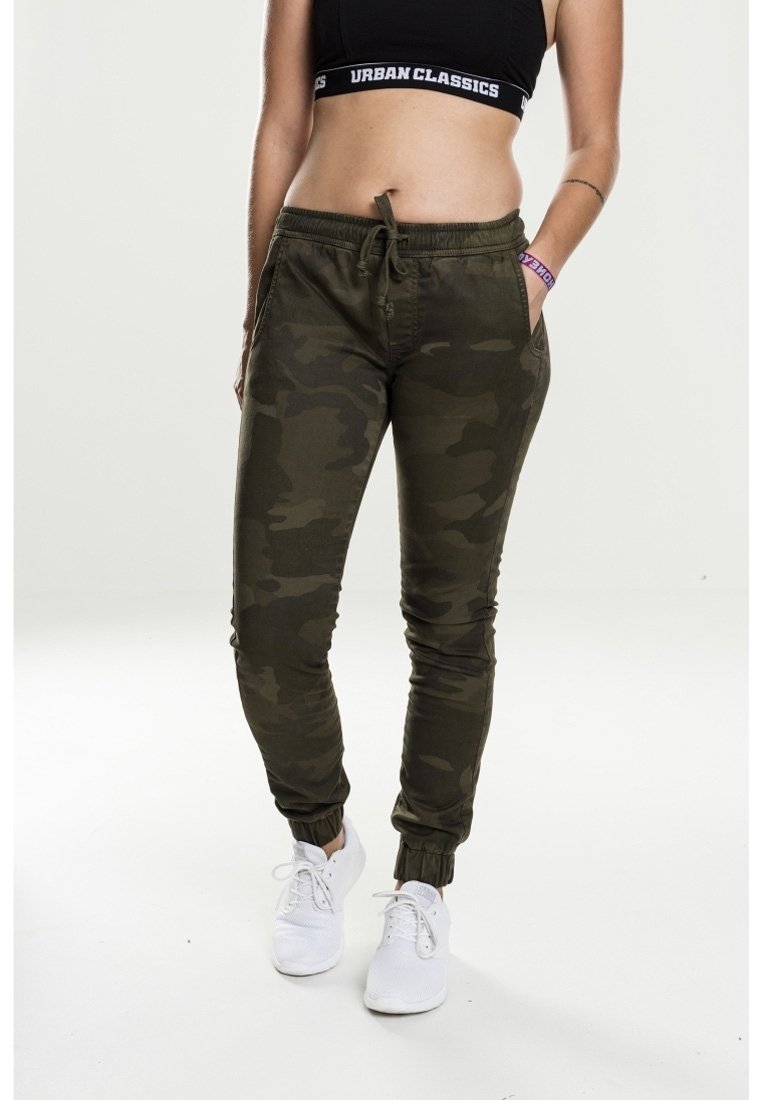 Femme portant un pantalon de jogging camouflage vert foncé, un crop top noir, des baskets blanches et un bracelet violet, debout, une main dans la poche.