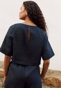 Navy blue cropped top with short sleeves, featuring a keyhole cutout at the back and textured fabric. Accented by a single button.