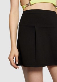 Svart plisserad kjol i mjukt tyg, med hög midja och sömnadsdetaljer. Modellen håller i nederkanten med ett avslappnat grepp.