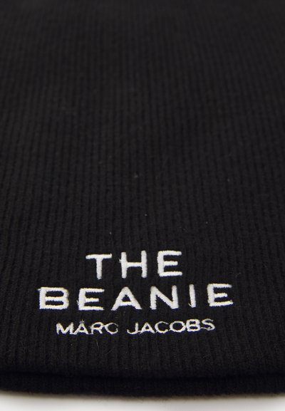 Black ribbed beanie with white embroidery displaying "THE BEANIE" and "MARC JACOBS" logos. Soft knit texture, close-up view.