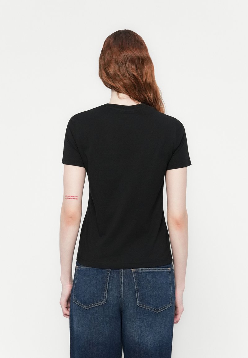 Persona con lunghi capelli rossi che indossa una maglietta nera e jeans blu, di spalle, mostrando un tatuaggio di testo rosso sul braccio sinistro.