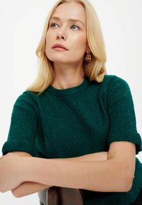 Maglione verde a maniche corte in morbido punto con scollatura rotonda. Le braccia del modello poggiano su una sedia di legno, mettendo in evidenza la trama e il colore del tessuto.