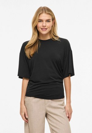 Fiatal nő hosszú, szőke hajjal, laza fekete rövid ujjú felsőt és bézs nadrágot viselve, fehér háttér előtt áll.