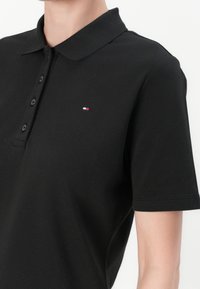 Personne portant un polo noir à manches courtes avec un petit logo rectangulaire rouge, blanc et bleu sur la poitrine gauche.