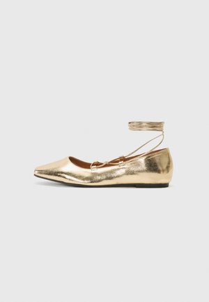 Guld flad balletsko med spids tå og rundremme om anklen på en ensartet lys baggrund.
