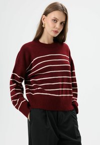 Bordeaux gebreide trui met horizontale crèmekleurige strepen, geribde ronde hals en manchetten; comfortabele pasvorm en zachte textuur. Zwarte broek zichtbaar.