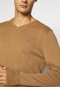 V-Ausschnitt Pullover aus leicht braunem Strickstoff mit langen Ärmeln und gerippten Bündchen. Kleiner Logo-Patch in Rot, Weiß und Blau auf der Brust.