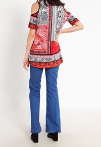 Túnica de hombros al descubierto con una mezcla de patrones rojos, negros y blancos; combinada con jeans de campana azules; presenta un diseño fluido y colores vibrantes.