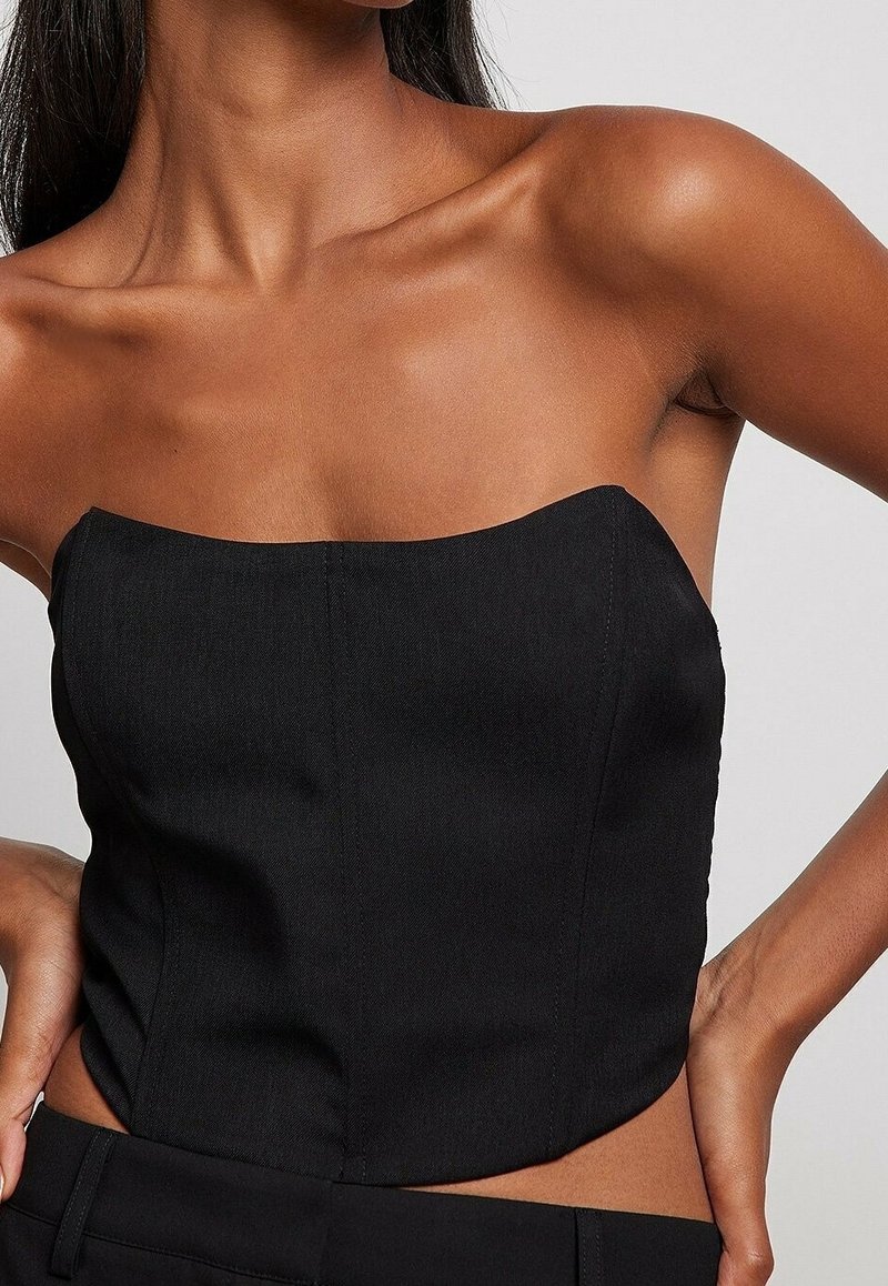 Femme portant un haut bustier noir avec un décolleté droit et un pantalon noir, les mains reposant sur ses hanches.