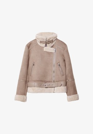 Vrouw draagt een beige shearling jas, wit overhemd, loszittende blauwe jeans en bruine suède schoenen, staand tegen een witte achtergrond.