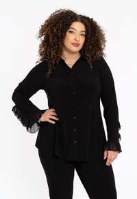 Zwarte blouse met een knoopsluiting aan de voorkant, lange mouwen en rimpelige doorschijnende accenten bij de manchetten. Gemaakt van een gladde stof met een vloeiende ontwerpen.