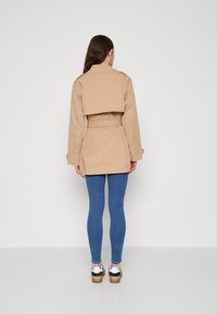 Beige trenchcoat met een hoge achterkant en een ceintuur in de taille, gecombineerd met blauwe skinny jeans en zwart-witte schoenen. Glad, gestructureerd materiaal.