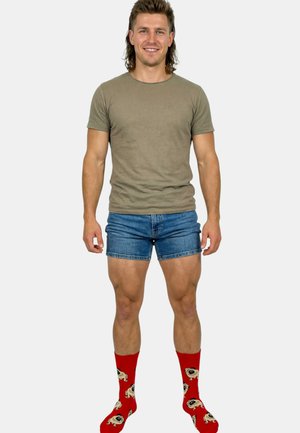 Jonge man staand, draagt een bruin t-shirt, korte blauwe denim shorts en rode sokken met cartoonachtige mopshondgezichten, glimlachend naar de camera.