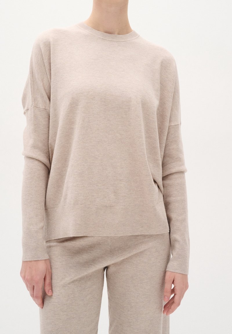 Personne portant un pull en maille beige à manches longues et un pantalon en maille assorti, debout devant un fond clair uni.