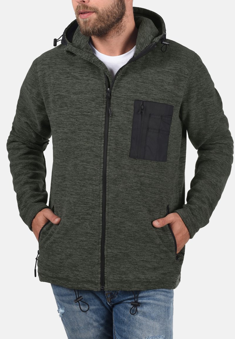 Chaqueta de forro polar de color verde oscuro con cremallera frontal, bolsillo en el pecho negro y capucha ajustable. Cuenta con bolsillos laterales y dobladillo con cordón.