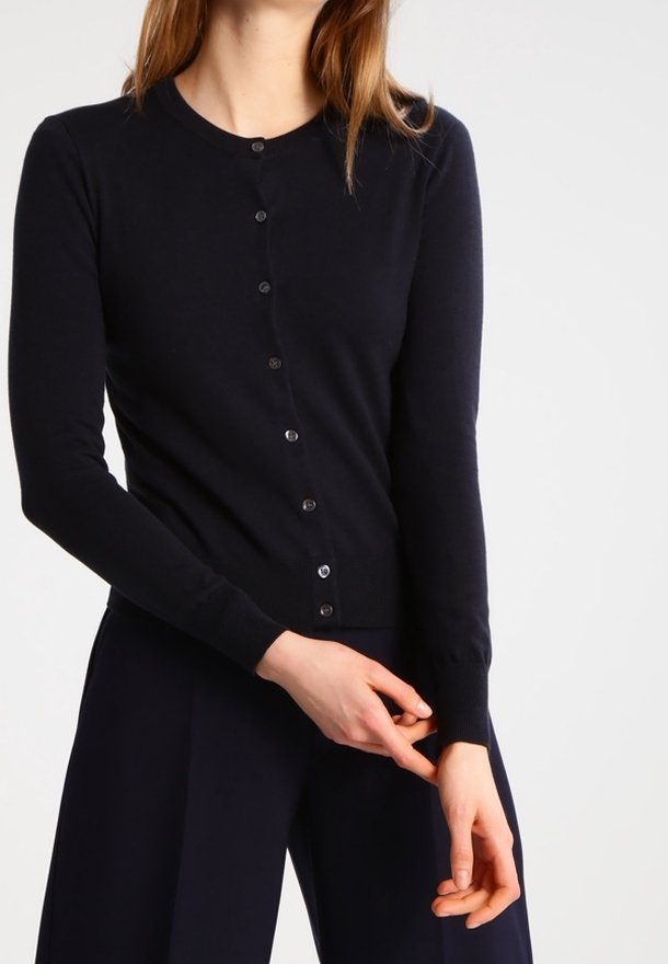 Kvinna iklädd en figurnära svart knappad cardigan och matchande svarta brallor, som justerar ärmen på sin vänstra arm.