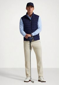 Námořnická prošívaná vesta přes světle modrou košili s dlouhým rukávem, béžové kalhoty a bílé golfové boty. Drží golfovou hůl, má na sobě tmavé rukavice a čepici.