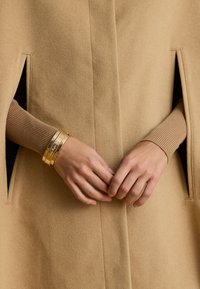 Μπεζ μάλλινο παλτό με πλαϊνές σχισμές, συνδυασμένο με καφέ ριχτό μανίκι. Τρία λεπτά χρυσά βραχιόλια στολισμένα στον καρπό, ανταν�ακλούν το φως.