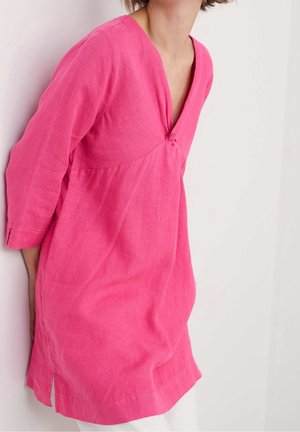 Kobieta w jaskraworóżowej tunice z długim rękawem, z dekoltem w serek i detalem w postaci supełka, stojąca na tle białej ściany.