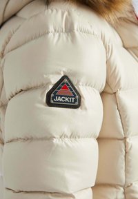 Beige pufferjack met een gewatteerd ontwerp. Heeft een driehoekige logo patch met blauwe, rode en witte accenten op de schouder.