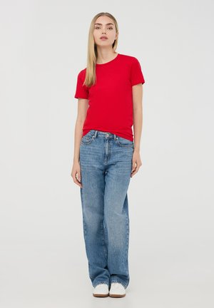 T-shirt rossa in cotone a maniche corte, abbinata a jeans in denim blu chiaro. Il modello indossa sneakers bianche, in piedi contro uno sfondo neutro.