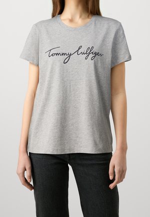 Kvinde iført en lysegrå kortærmet T-shirt med sort "Tommy Hilfiger" skrift over brystet og mørke bukser.