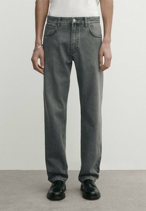 Homme portant un jean gris ample, un t-shirt blanc, des chaussures noires et un bracelet en argent, debout sur un sol gris avec un fond uni.