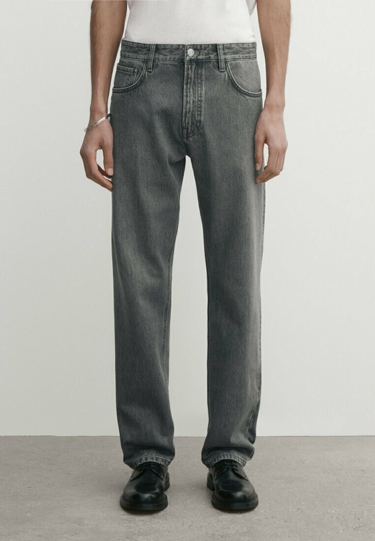 Homme portant un jean gris ample, un t-shirt blanc, des chaussures noires et un bracelet en argent, debout sur un sol gris avec un fond uni.