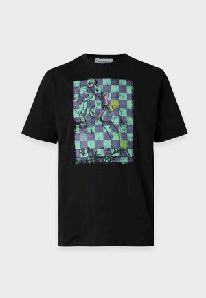 Junger Mann mit schwarzem Grafik-T-Shirt, dunkelgrauen Jeans und weißen Turnschuhen, der vor einem einfarbigen hellen Hintergrund steht.