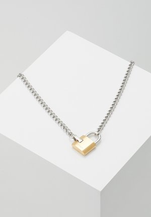 Collier en chaîne argentée avec un petit pendentif en forme de cadenas en or, posé sur un cube blanc sur un fond gris.