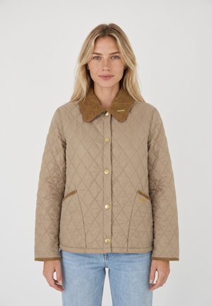 Vrouw draagt een beige doorgestikte jas met een bruine kraag en afwerking, gouden knopen en lichtblauwe spijkerbroek tegen een effen witte achtergrond.