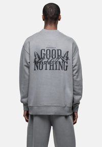 Felpa grigia con maniche lunghe, caratterizzata da una grande grafica nera sul retro con il testo "GOOD FOR NOTHING" e illustrazioni di farfalle.