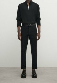 Polo noir à manches longues avec col zippé, associé à un pantalon noir et des chaussures à lacets, présentant une coupe ajustée et une texture lisse.