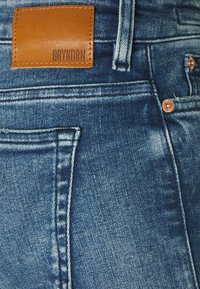 Modré džínové kalhoty s hnědou koženou etiketou s vyraženým nápisem "DRYKORN", viditelným šitím a měděnými nýty.