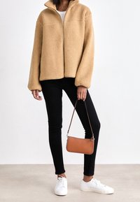 Woman wearing beige fuzzy zip-up jacket, black slim pants, white sneakers, holding small brown handbag by strap against plain background.