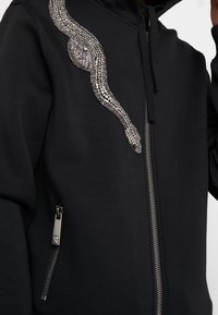 Sudadera con cremallera negra que presenta un adorno metálico en forma de serpiente en el lado izquierdo y un bolsillo con cremallera en la parte inferior derecha.