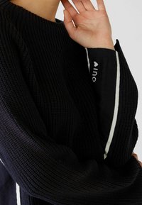 Pull en maille noire avec un col rond, agrémenté d'accents à rayures blanches et d'un détail textuel sur la manche. Tissu texturé avec une coupe ample.