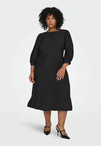 Melna kleita līdz ceļgaliem ar brīvu piegriezumu, pūkainām piedurknēm un plašu dekoltē. Izgatavota no teksturēta auduma, kombinēta ar norādītām melnām kurpēm ar smailiem purngaliem.