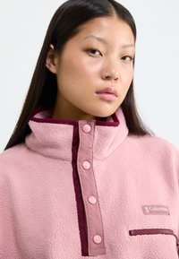 Pull en polaire rose clair, avec un col montant, des accents bordeaux, des boutons à l'avant et une poche zippée. Texture douce avec un détail de logo.