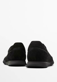 Scarpe sportive in suede nero con una texture liscia, design a profilo basso e suole in gomma nera, viste da dietro. Stile minimalista.