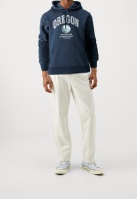 Námořnická modrá mikina s potiskem "Národní park Crater Lake v Oregonu", klokaní kapsa, v kombinaci s světlými béžovými kalhotami a modrými teniskami.