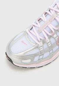 Vit och silver nät-sneaker med rosa detaljer, rosa snörning och en liten rosa Nike-swoosh nära tån.