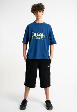 Nastolatek stoi zwrócony do przodu, ubrany w niebieską koszulkę z napisem „REAL Certified”, czarne krótkie spodenki do kolan, białe skarpetki i czarne sneakersy.