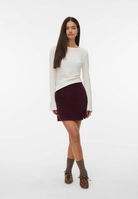 Eine Frau mit langen, glatten Haaren trägt ein weißes, strukturiertes Langarmoberteil, einen weinroten Minirock mit Schlitz, braune Socken und braune Schuhe und steht klassisch mit übergeschlagenen Beinen.