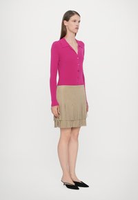 Cardigan roz cu mânecă lungă și nasturi, purtat cu o fustă din suede bej cu franjuri. Modelul poartă mule negre cu toc, stând pe un fundal simplu.