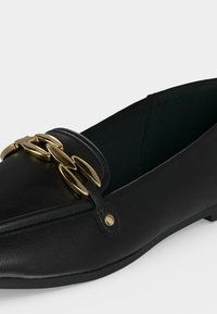 Mocassin en cuir noir avec bout pointu et chaîne décorative dorée sur le dessus.