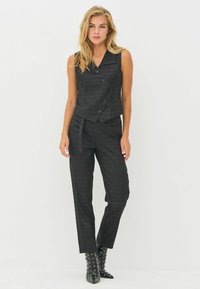 Zwart, textuurvol tweedelig outfit bestaande uit een mouwloze, dubbelgeknoopte vest en strakke broeken met een vastgebonden taille. Met gespijkerde zwarte laarzen wordt de look compleet.