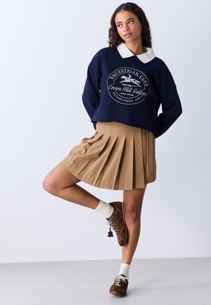 Jeune femme debout sur une jambe, portant un sweat bleu marine avec un col blanc, une jupe plissée beige, des chaussettes blanches et des chaussures à motif léopard.